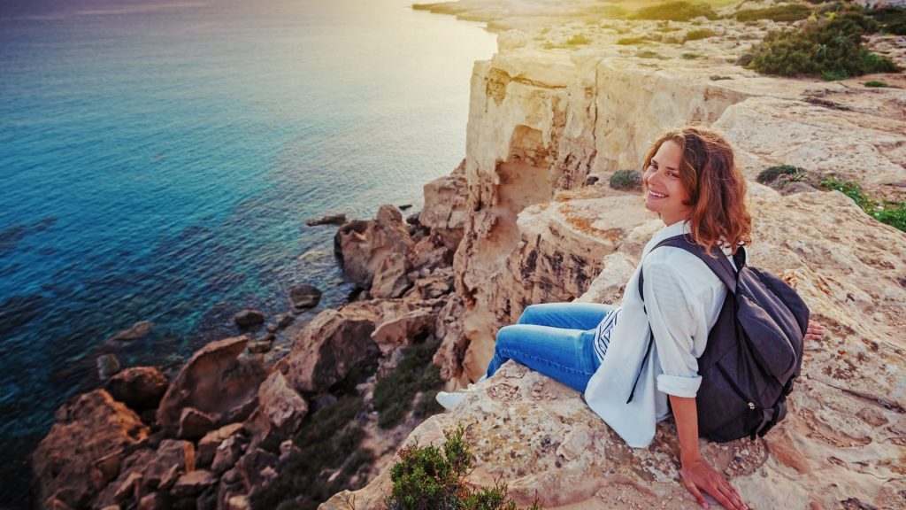 A stylish young woman traveler watches a beautiful sunset on the rocks on the beach, Cyprus, Cape Greco, a popular destination for summer travel in Europe