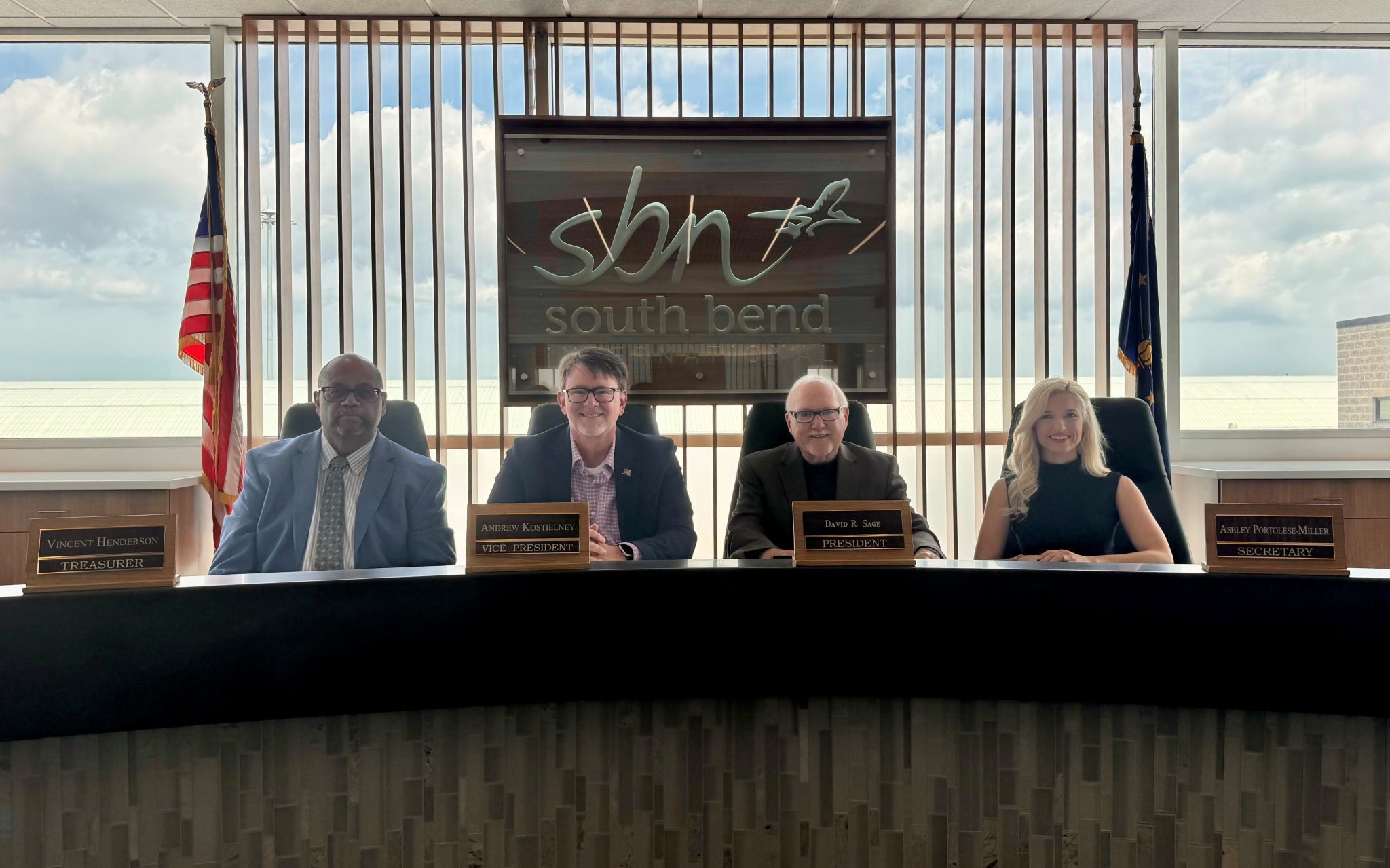 Photo of the Board of St. Joseph County Airport Authority seated with their name plates. A giant sign with the SBN logo is behind them with two flags to the left and right. 