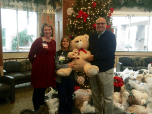 There are three people in the photo, two nurses and the one in the middle is holding a teddy bear. On the other side of the teddy bear is Mike Daigle, CEO. There are bags of bears piled in the foreground of the photo with the background being a hospital lobby.
