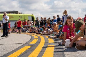 This is a photo of children sitting in a half circle on pavement. There is an ARFF truck in the background to the left. The fire chief is talking on his radio in front of the ARFF truck. 