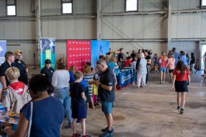 This is a photo inside an airport hangar where there are booths and backdrops set up for a career fair. In the foreground, people are milling about at the booths. 