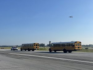 This is a photo of a runway at South Bend International Airport. There is an operations vehicle to the far left leading two school buses down the runway. A small plane is flying in the sky overhead. 