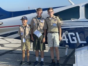 Three boy scouts are standing in the center of the photo smiling in front of a small plane that is in the background. 