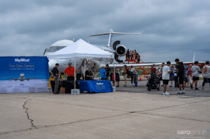 In the middle ground of the photo, there is a big backdrop for SkyWest with a tent and table booth. There is a plane behind it, barely seen, and people are milling about by the booth and in front of the plane. 
